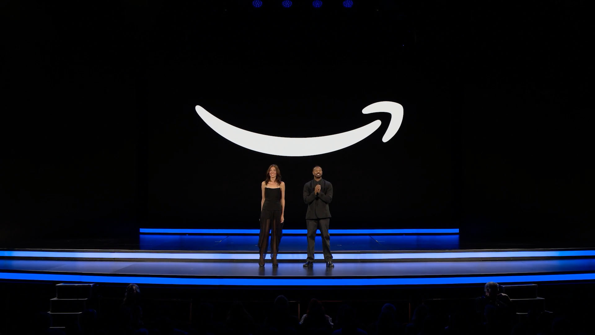 Two presenters on a dark stage beneath a large white Amazon smile.
